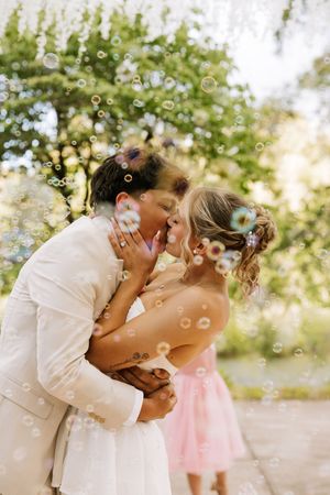 Couple sharing a romantic kiss surrounded by floating bubbles outdoors, with flowers in their hair on a wedding day.