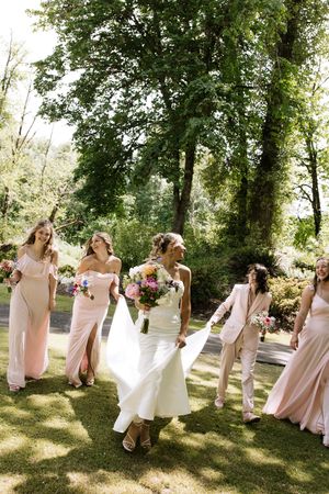 Bride and bridesmaids in blush pink dresses walk together through a lush green garden setting.