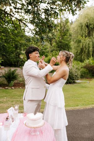 Two brides feed each other wedding cake outdoors in a lush garden setting, wearing white dress and beige suit.