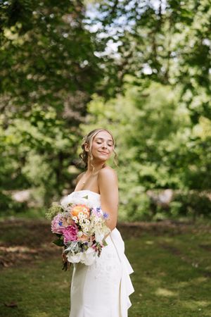 Bride in white strapless gown holding colorful bouquet with pink and purple flowers, smiling outdoors in lush green garden.
