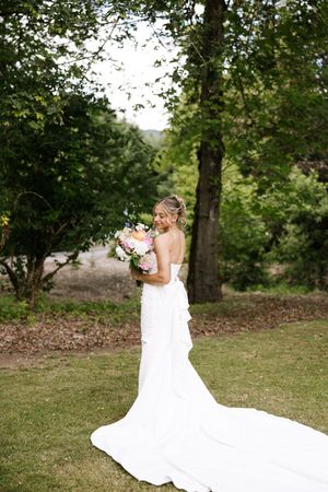 Elegant bride in strapless white gown with train holds colorful bouquet in a lush garden setting.