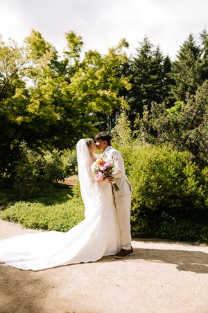 Bride and groom kissing in a lush garden, bride wearing a flowing white gown with veil holding a pink bouquet.
