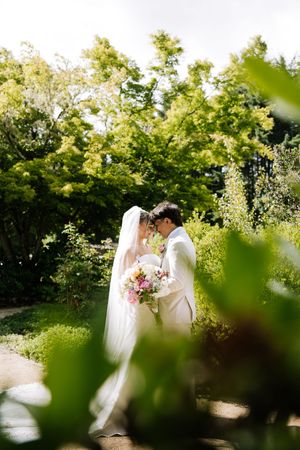 Newlyweds touch foreheads tenderly in a sunlit garden, bride holding a vibrant bouquet in a white gown.