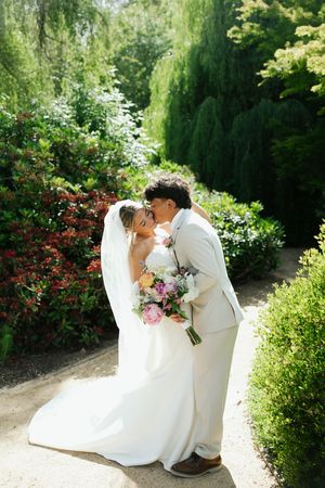 Groom kisses bride on cheek in a sunny garden path, bride holding a pink and white floral bouquet.