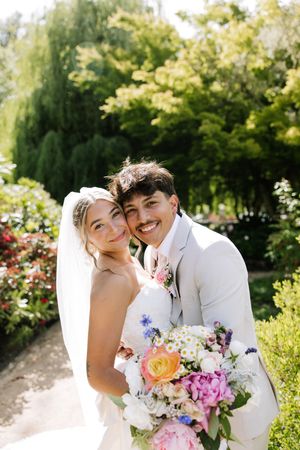 Smiling bride and groom pose together in a sunny garden, bride holding a colorful bouquet of pink and white flowers.