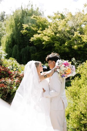 Bride in white gown and veil holds colorful bouquet while embracing groom in cream suit outdoors.