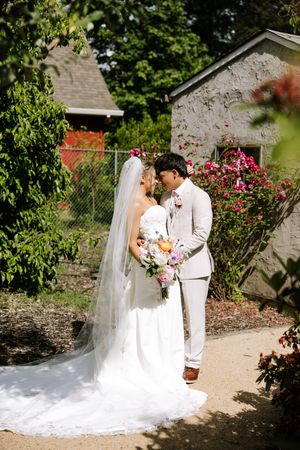 Bride and groom share a romantic nose-to-nose moment near blooming flowers outside a stone building.