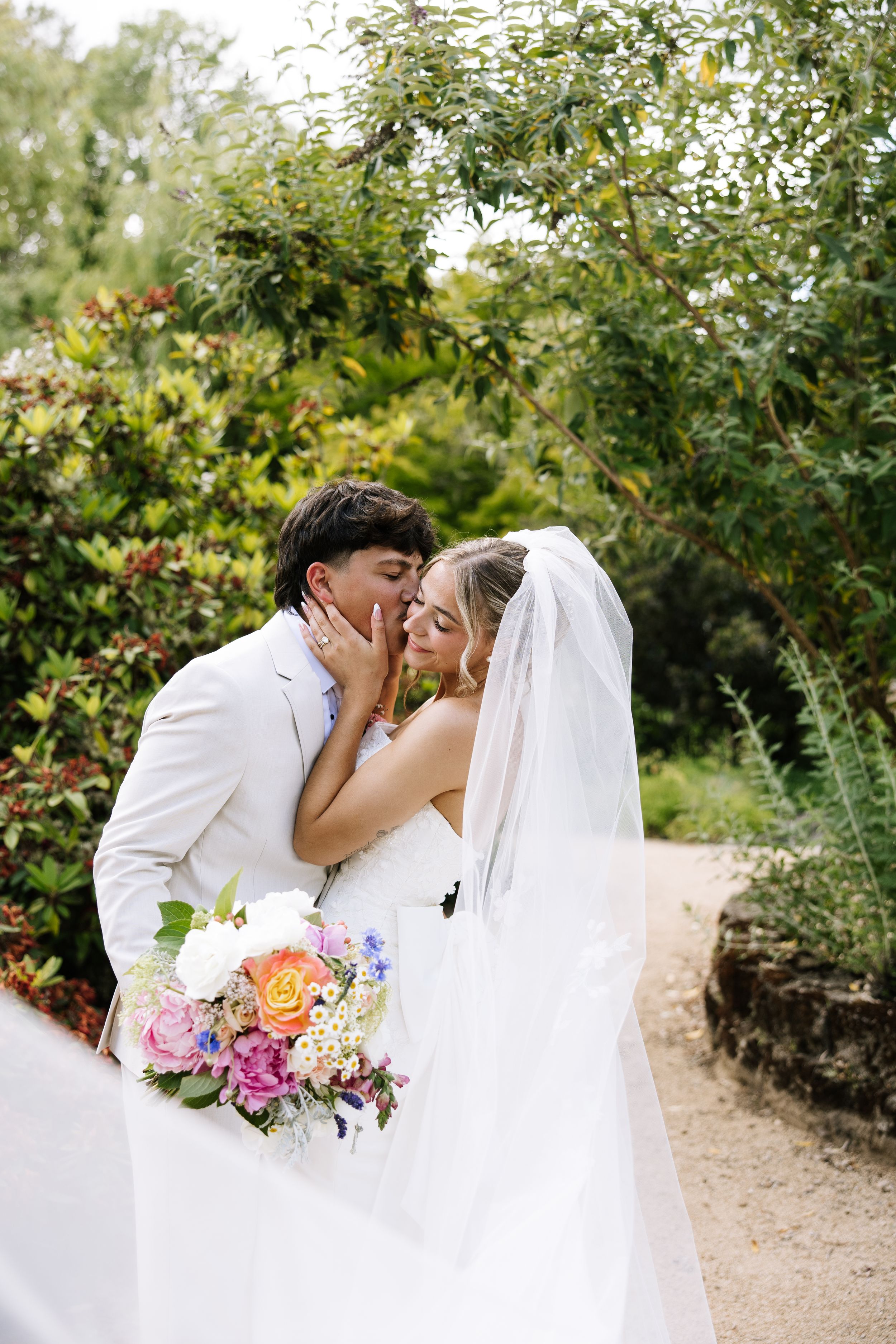 Bride and groom share a romantic kiss in a lush garden, bride holding a colorful bouquet with veil flowing.