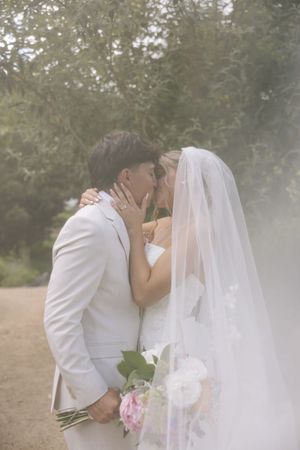 Couple sharing a romantic kiss surrounded by soft mist, bride in white gown with veil holding pink flowers.