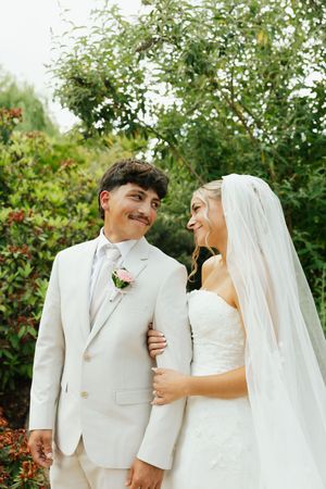 Happy bride and groom walk together outdoors, bride in lace dress with veil, groom in light beige suit with pink boutonniere.