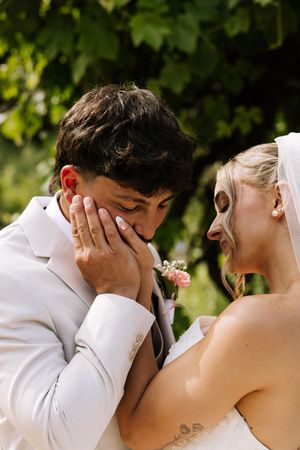Emotional groom in white suit covers his face as bride in veil smiles tenderly in garden setting.