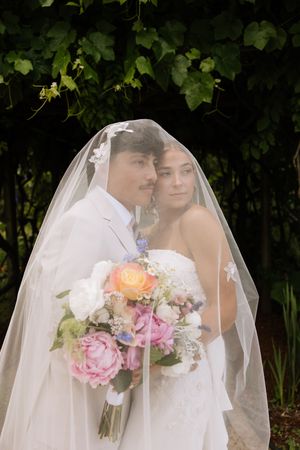 Newlyweds posing together under a sheer wedding veil with a vibrant floral bouquet in a lush garden setting.