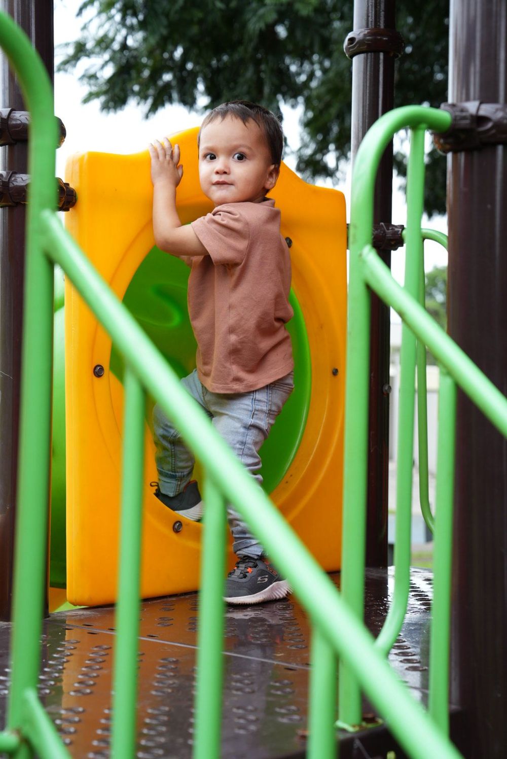 Cute toddler boy in brown shirt playing on colorful yellow and green playground equipment.
