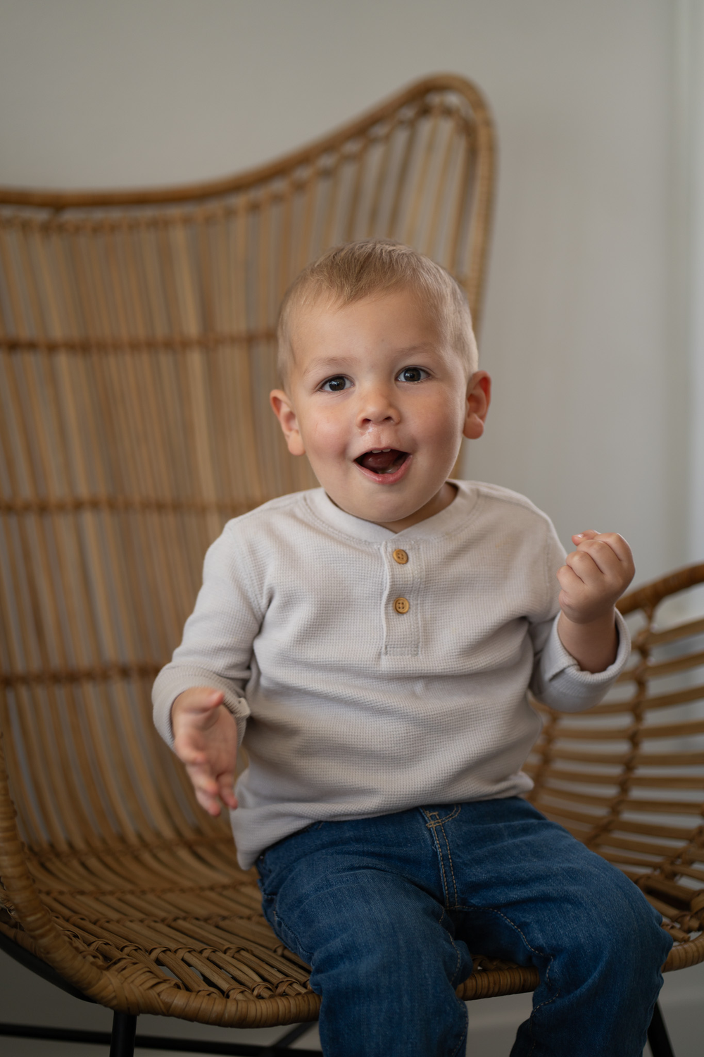 Happy toddler boy in gray henley shirt sitting in wicker rattan chair laughing.