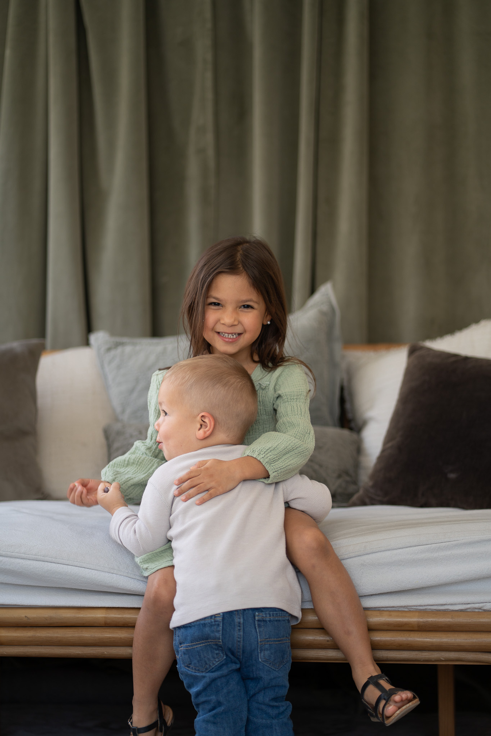 Smiling girl in green dress hugging toddler brother while sitting on bed with pillows.
