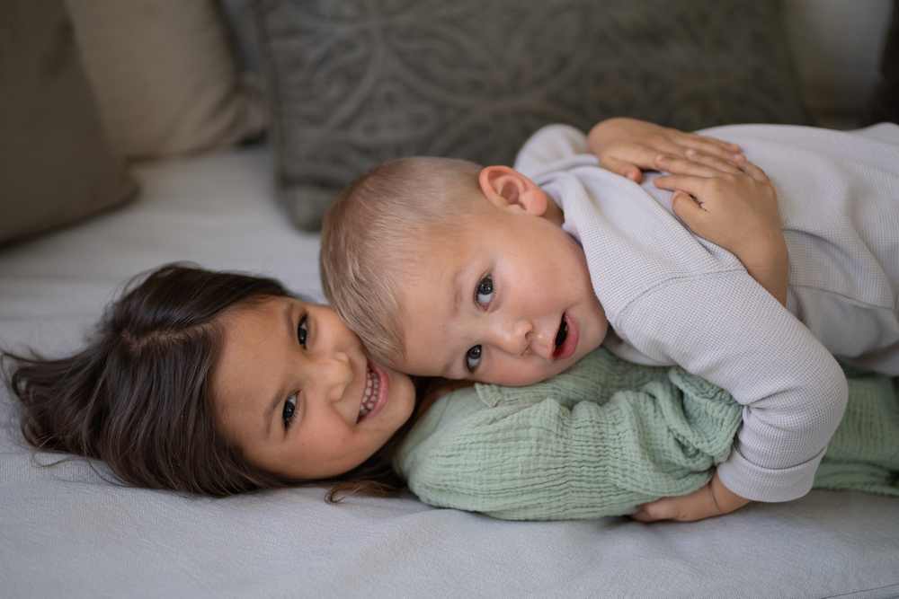 Young girl lying on bed smiling while holding baby sibling on her back.