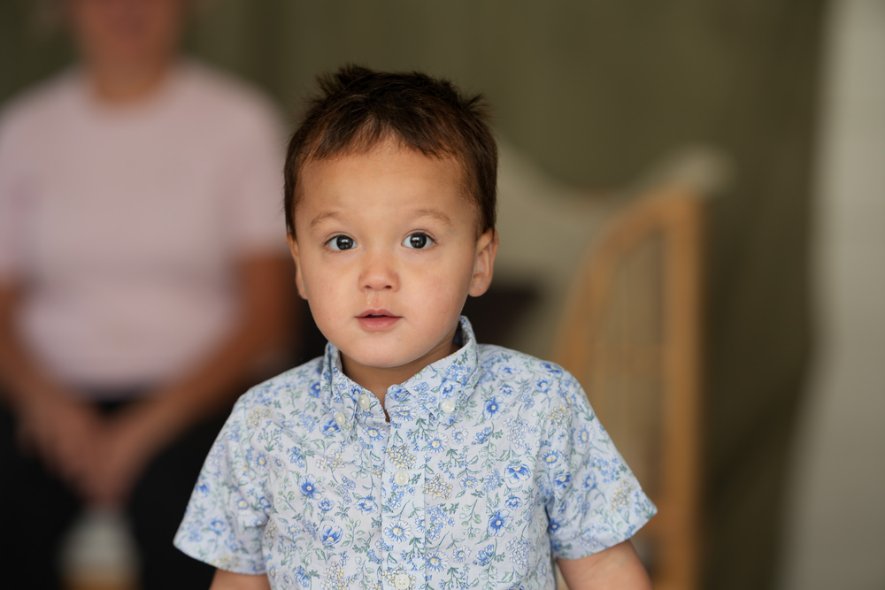 Cute toddler boy in blue floral shirt looking at camera with blurred background.