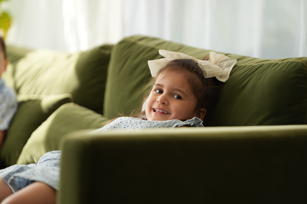 Cheerful little girl with bow in hair peeking over back of green sofa.