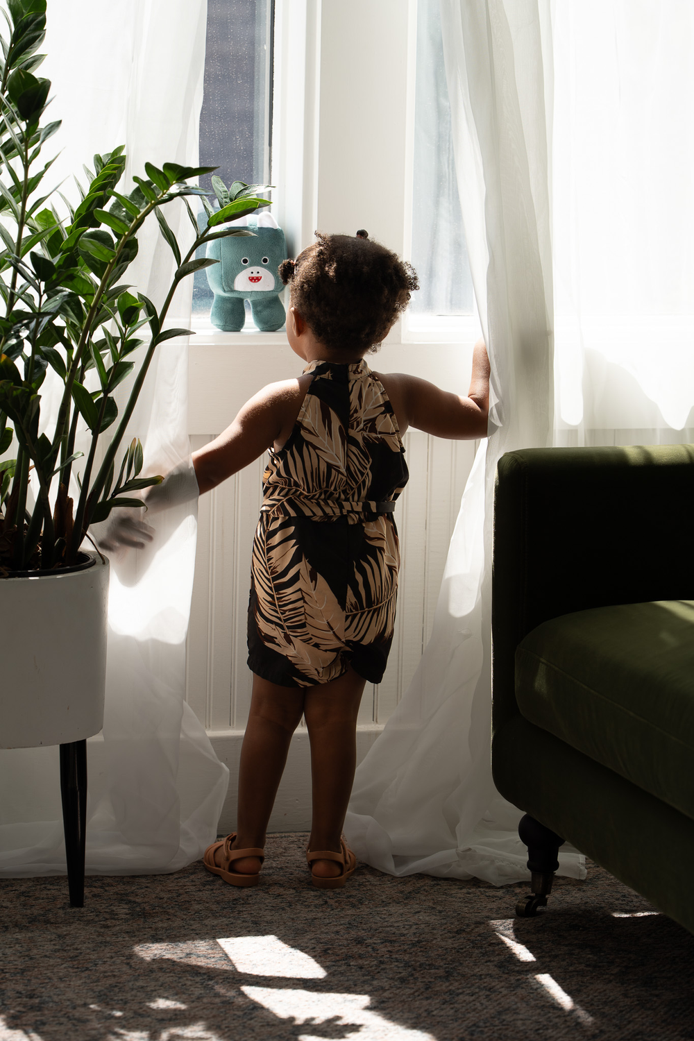 Toddler girl in tropical romper standing at window beside houseplant and stuffed bear.
