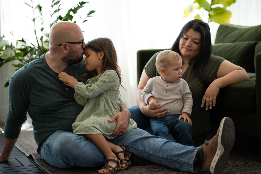 Family of four sitting on floor together with two children and green couch backdrop.