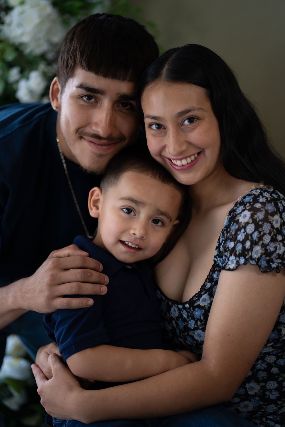 Young family portrait with parents embracing smiling toddler boy against floral background.
