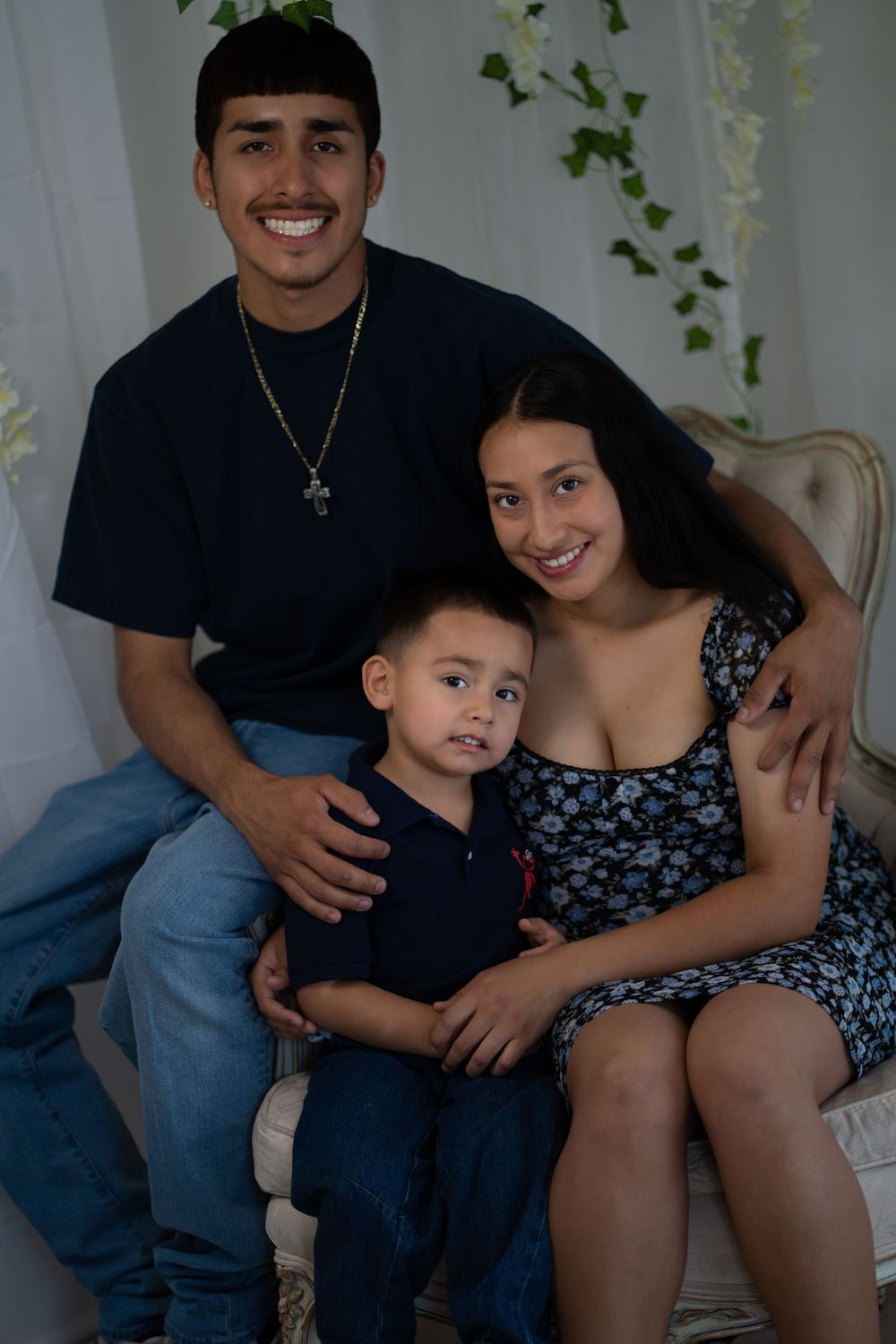 Young Latino family of three smiling together in a bright white studio portrait setting.