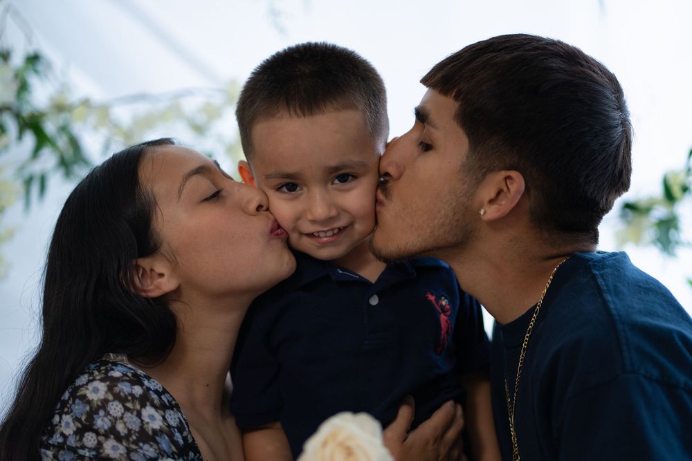 Loving parents kissing their young son on both cheeks simultaneously indoors.