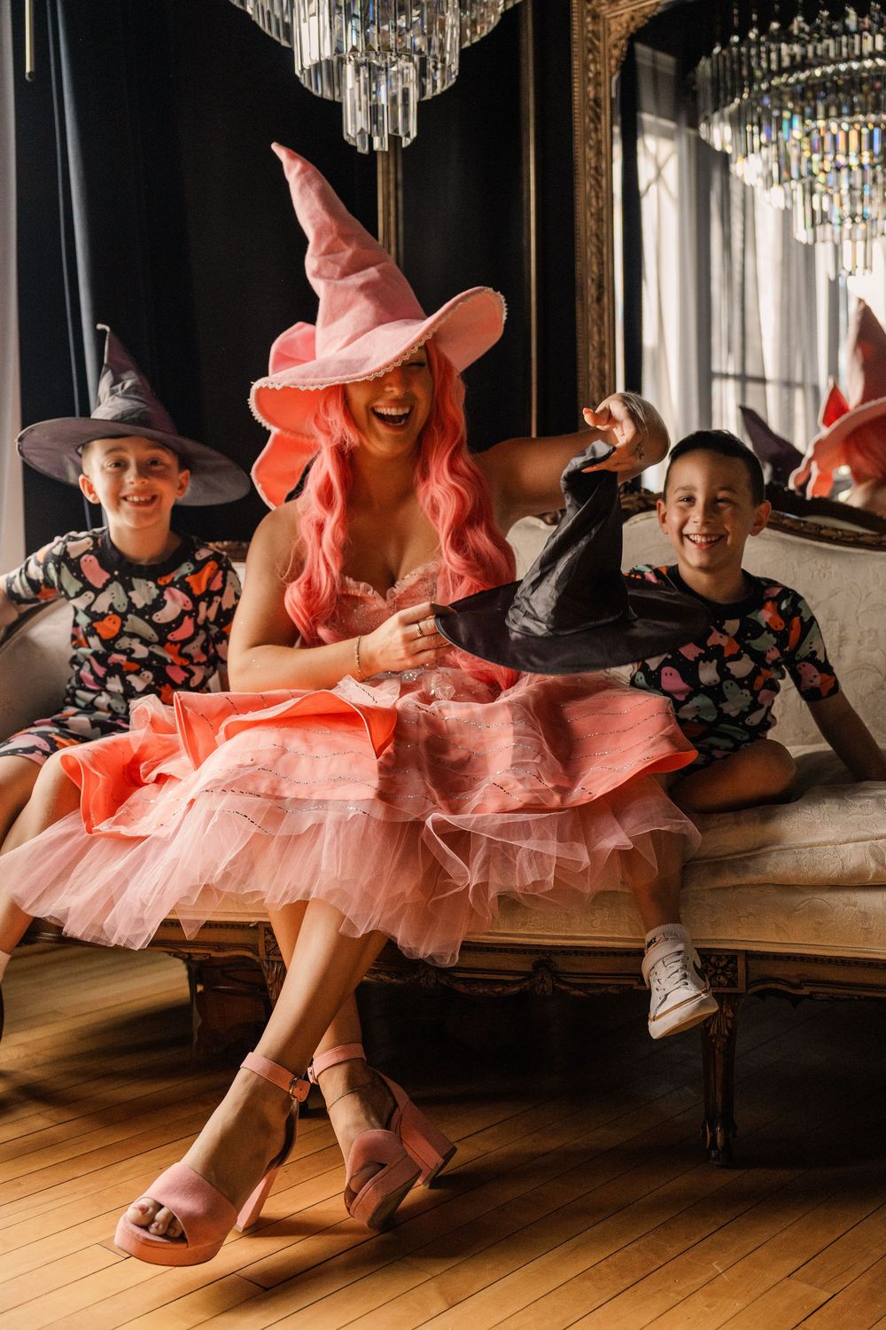 Woman in pink witch costume laughing with two children in Halloween costumes on ornate sofa.