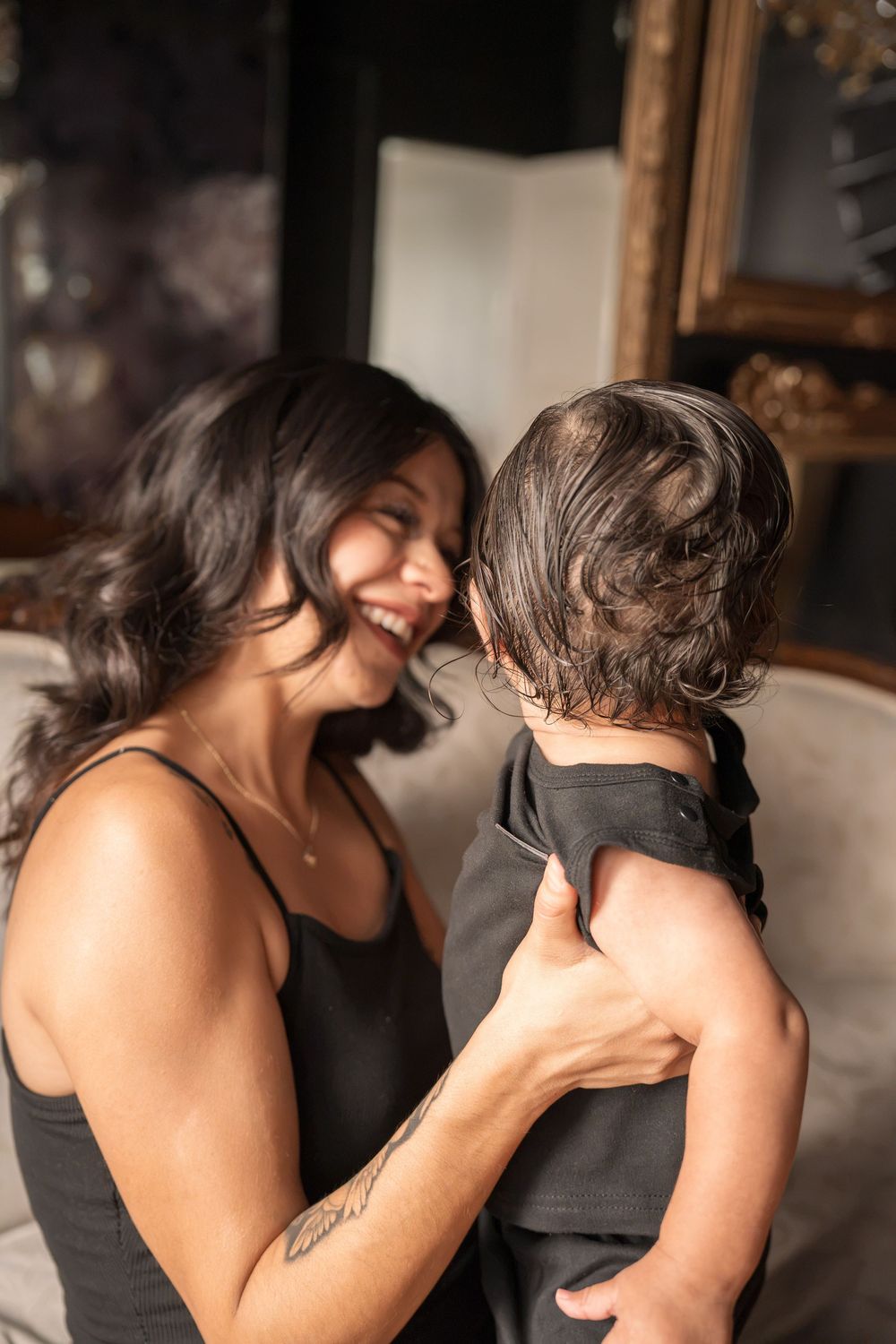 Joyful dark-haired woman laughing while holding a young child in a warmly lit room.