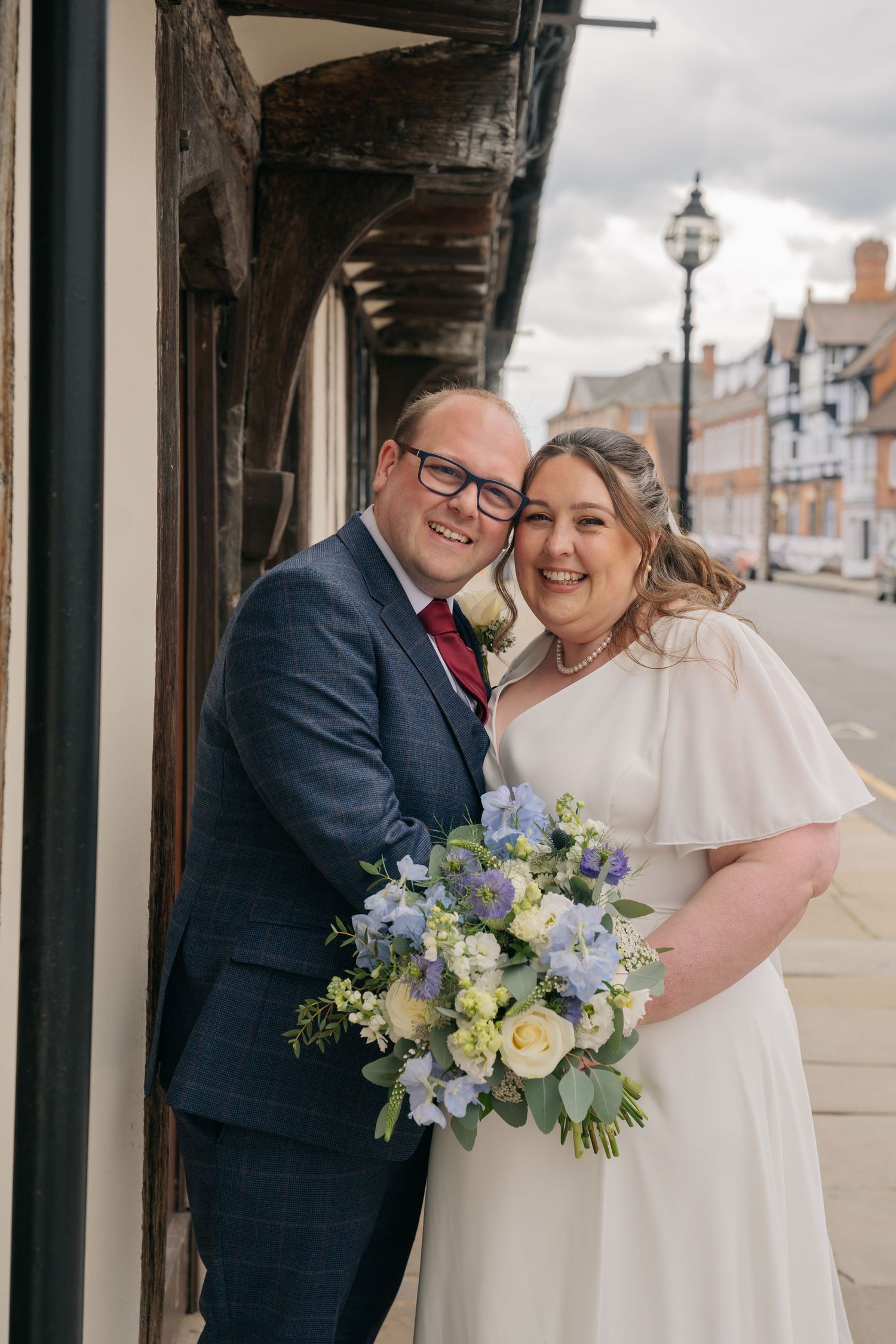 Smiling newlyweds pose outdoors, bride in white cape dress holding blue and white bouquet, groom in navy suit with red tie.
