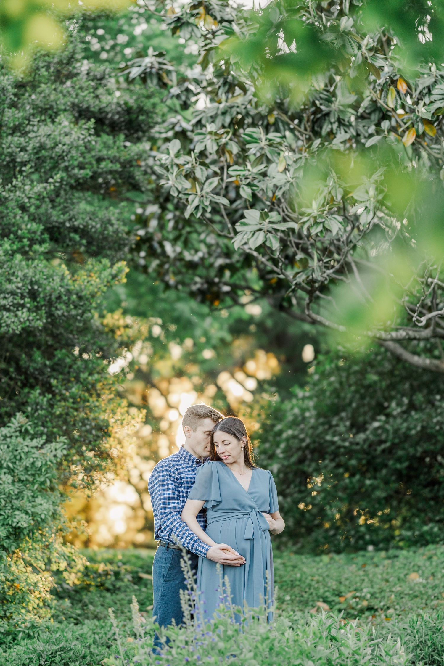 Couple sharing a tender maternity moment outdoors, surrounded by lush green trees in soft golden light.