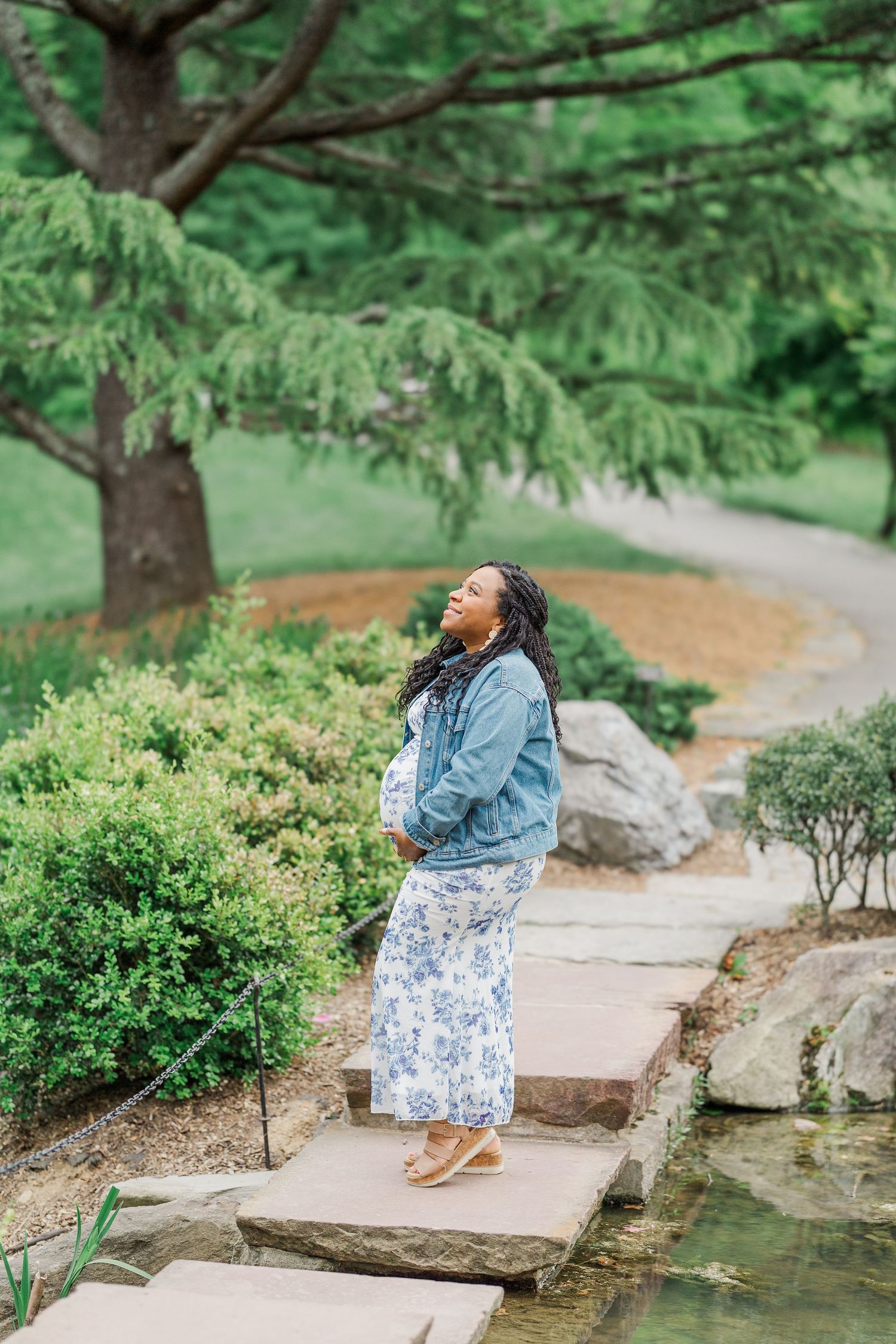 Pregnant woman in denim jacket and floral skirt standing on stepping stones in a lush green garden.