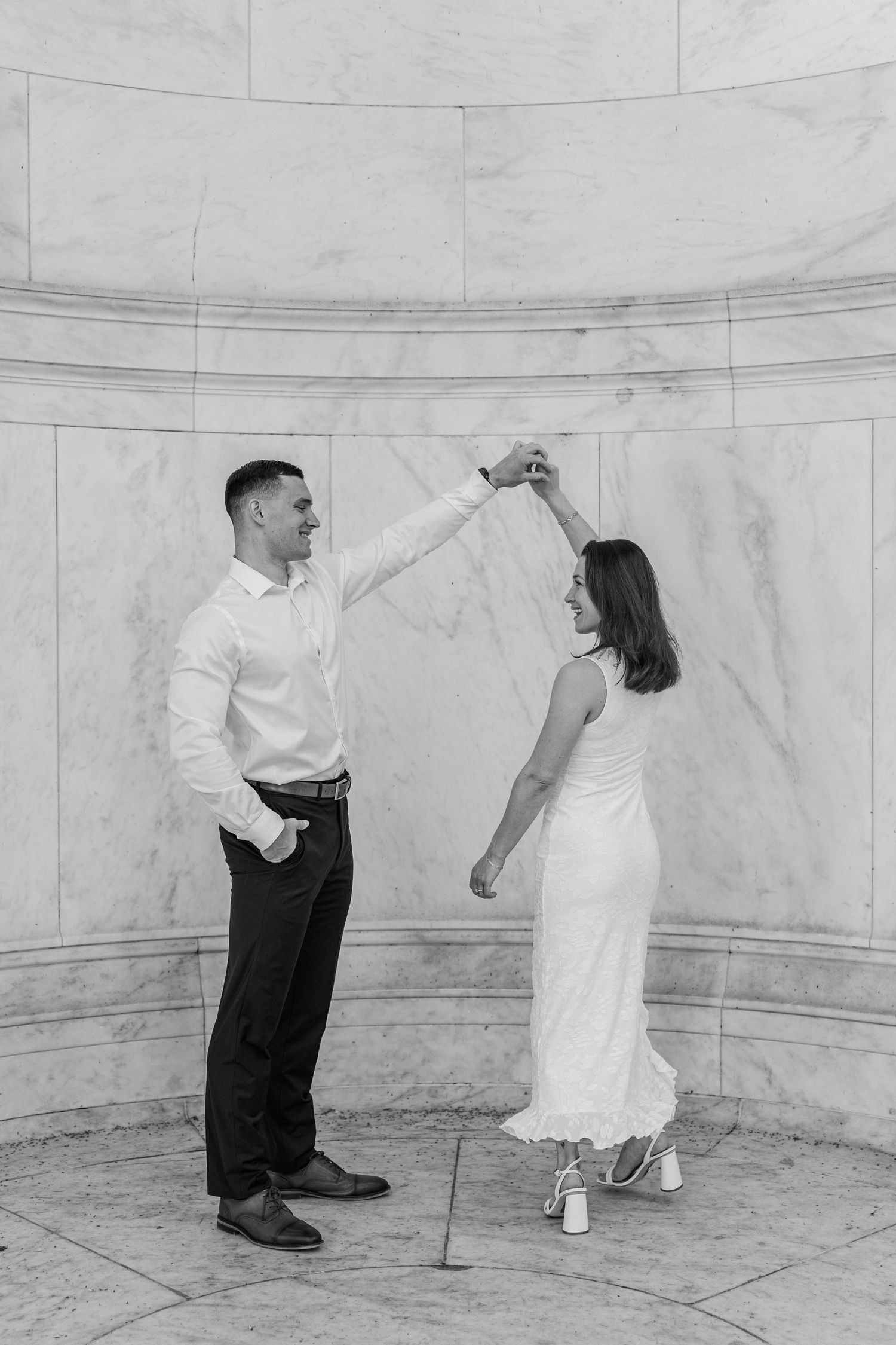 Black and white photo of a couple dancing elegantly in front of marble columns, man spinning woman in white dress.