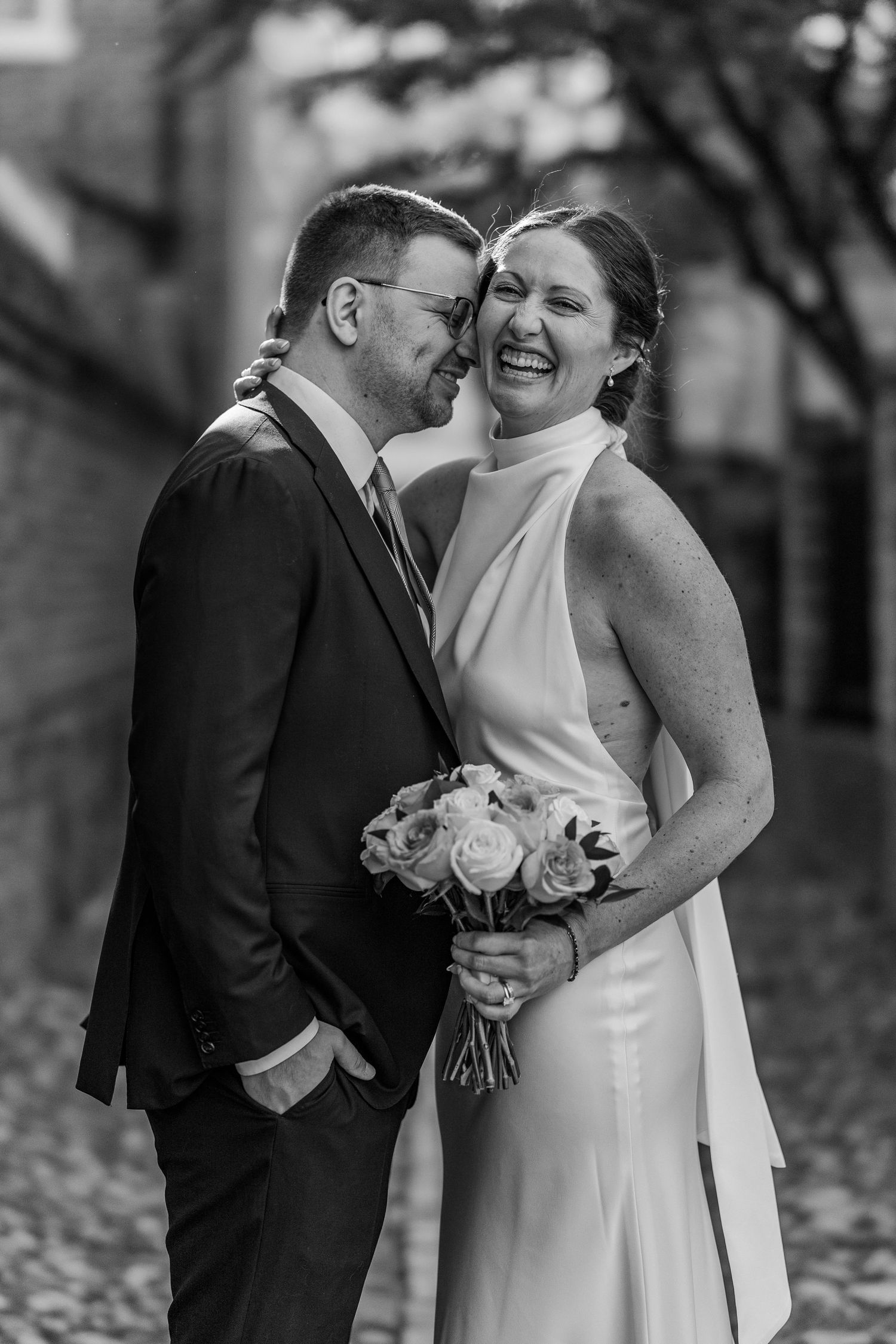 Black and white wedding photo of smiling bride holding bouquet and groom in suit sharing a joyful moment outdoors.