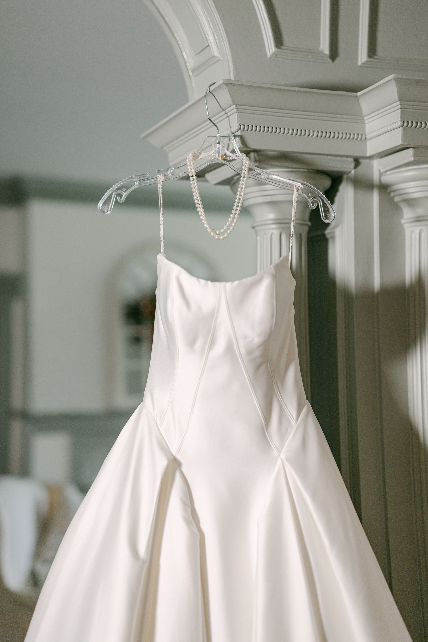 An elegant white satin strapless wedding gown hangs on a pearl hanger from a decorative mantelpiece.