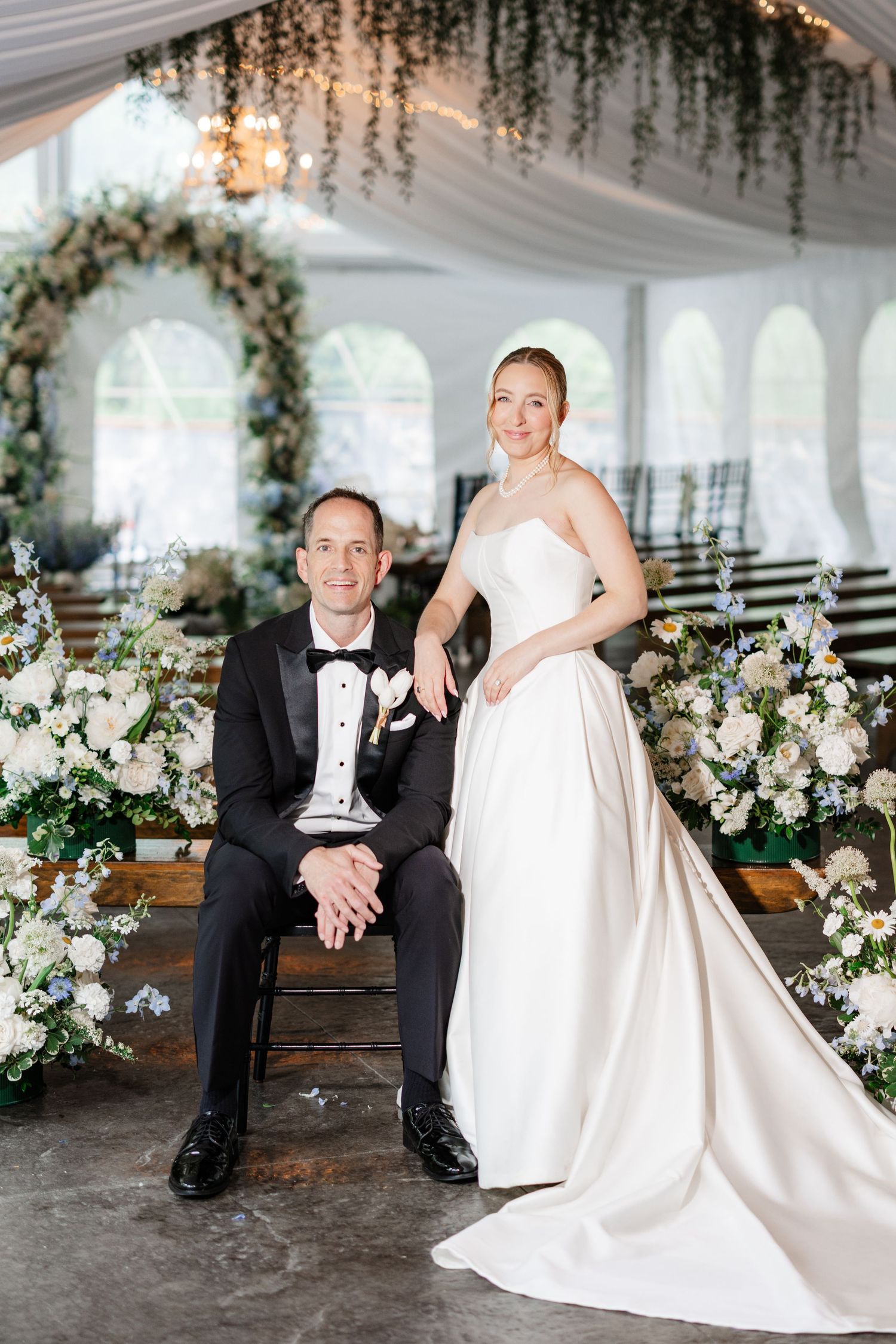Bride in strapless white ball gown standing beside seated groom in tuxedo inside floral decorated wedding tent.