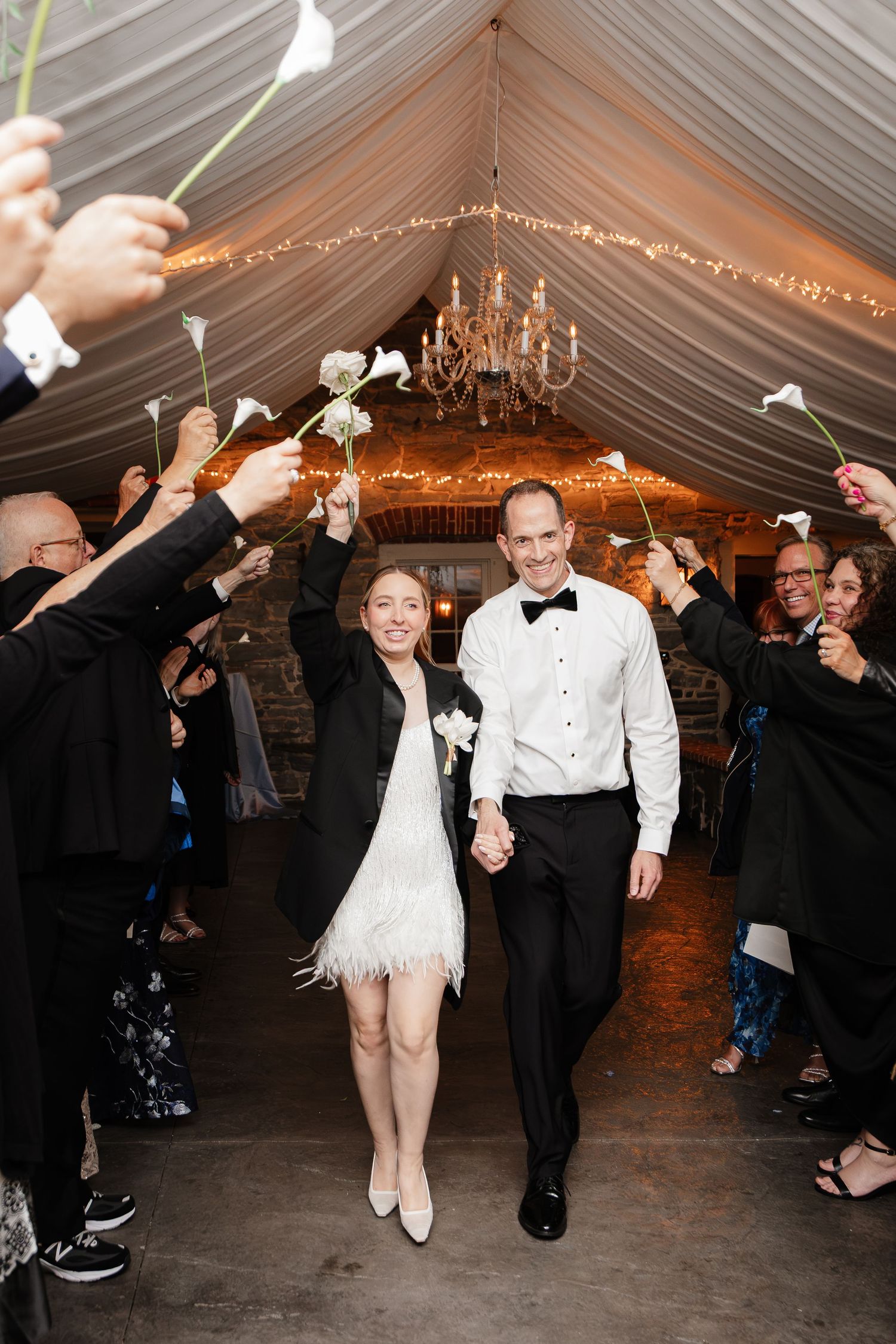 Bride and groom walk through calla lily sendoff under chandelier in elegant tented wedding reception.