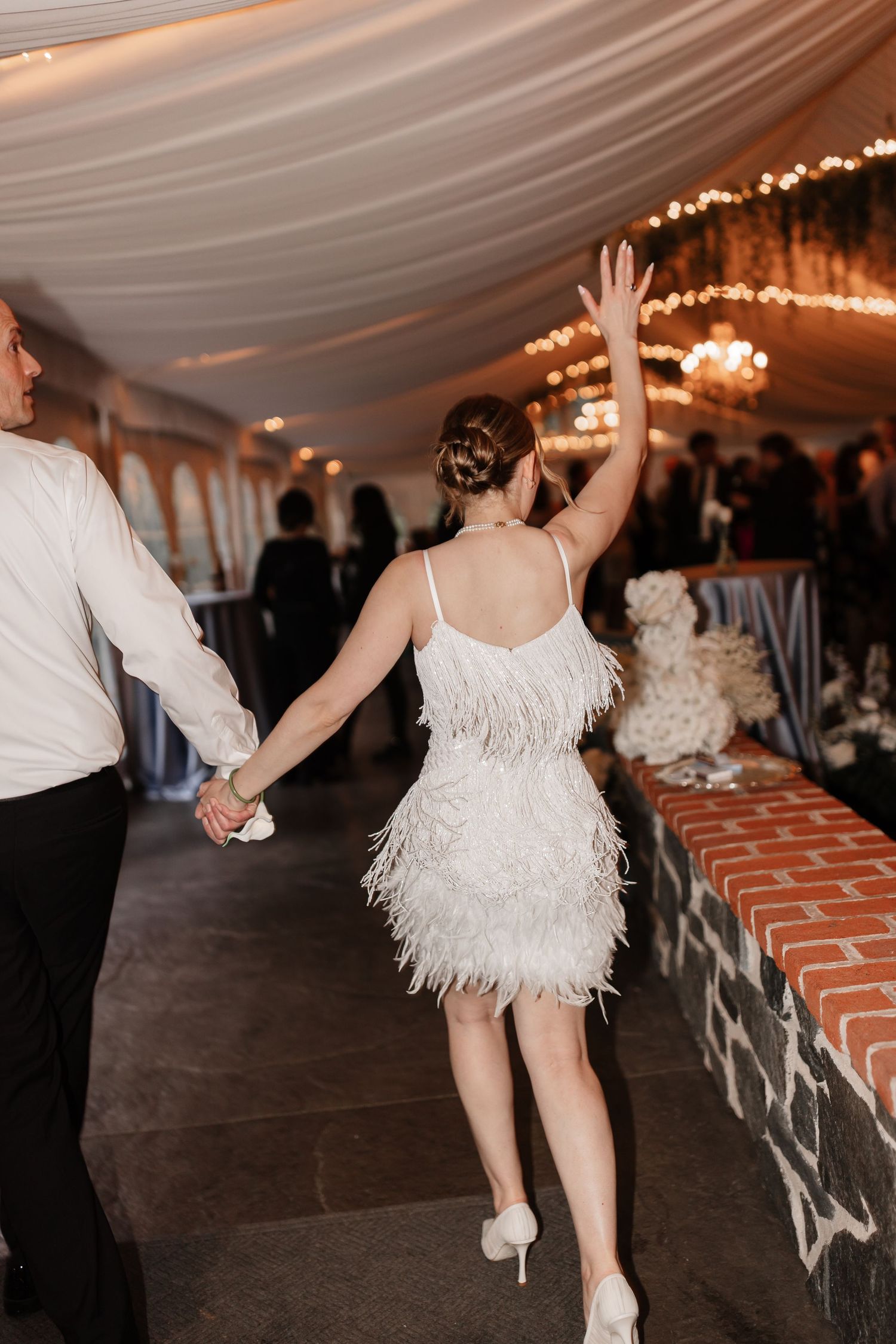 Bride in white feather mini dress holds hand of groom while raising arm at tented wedding reception.