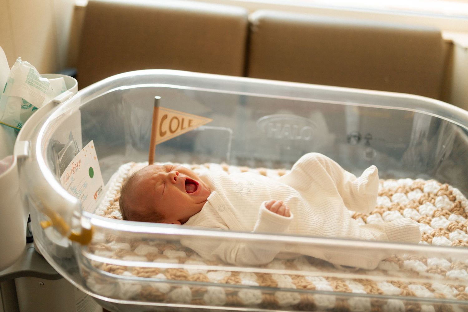 Newborn baby yawning in a clear hospital bassinet with a small personalized flag, swaddled in white at University of Tennessee Medical Cente