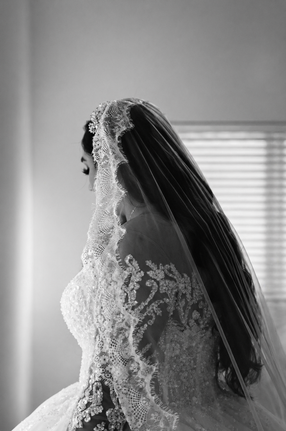 Black and white bridal portrait of a bride in lace wedding gown and veil, standing by a window.