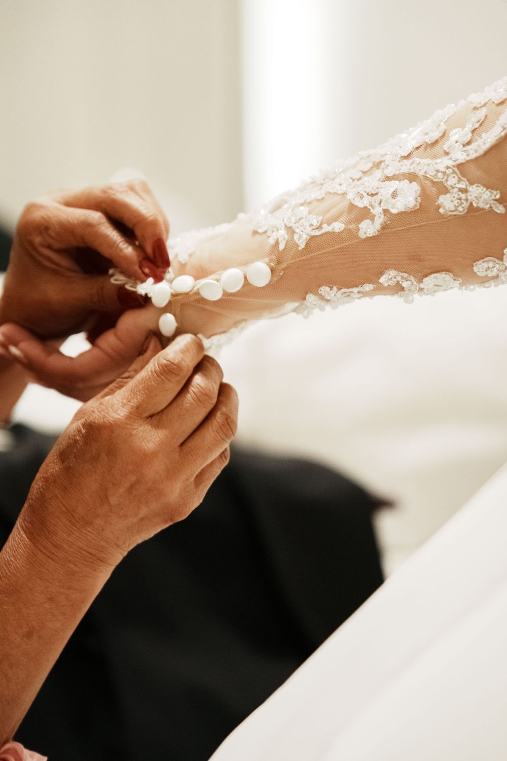 Close-up of hands fastening pearl buttons on a lace wedding dress sleeve.