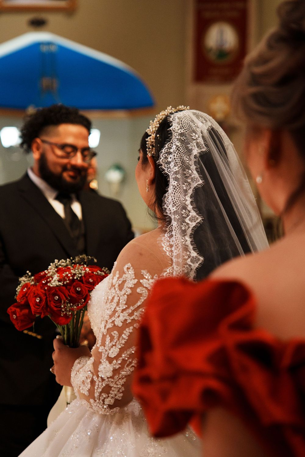 Bride in lace wedding dress holding red rose bouquet faces groom in black suit during church wedding ceremony.