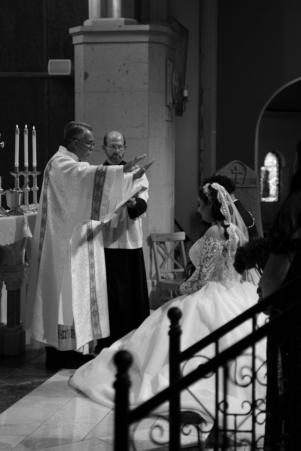 Priest blessing bride in white gown during Catholic wedding ceremony inside a church, black and white photo.