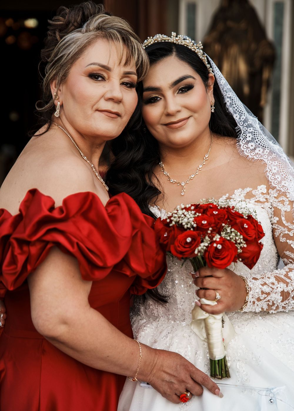 Bride in white lace gown with red rose bouquet poses with mother in red off-shoulder dress.