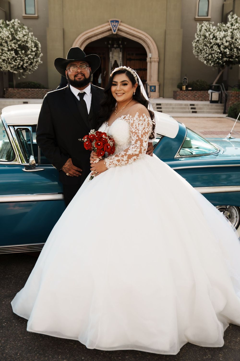 Bride in a ball gown and groom in a black cowboy hat pose beside a vintage teal car outside a church.