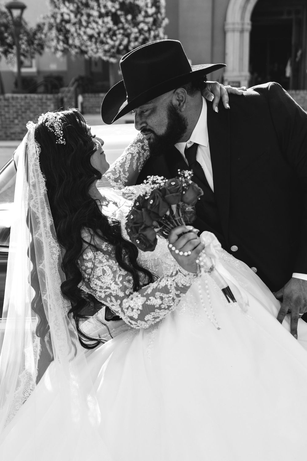 Bride and groom share a romantic moment beside a vintage car in black and white wedding photography.