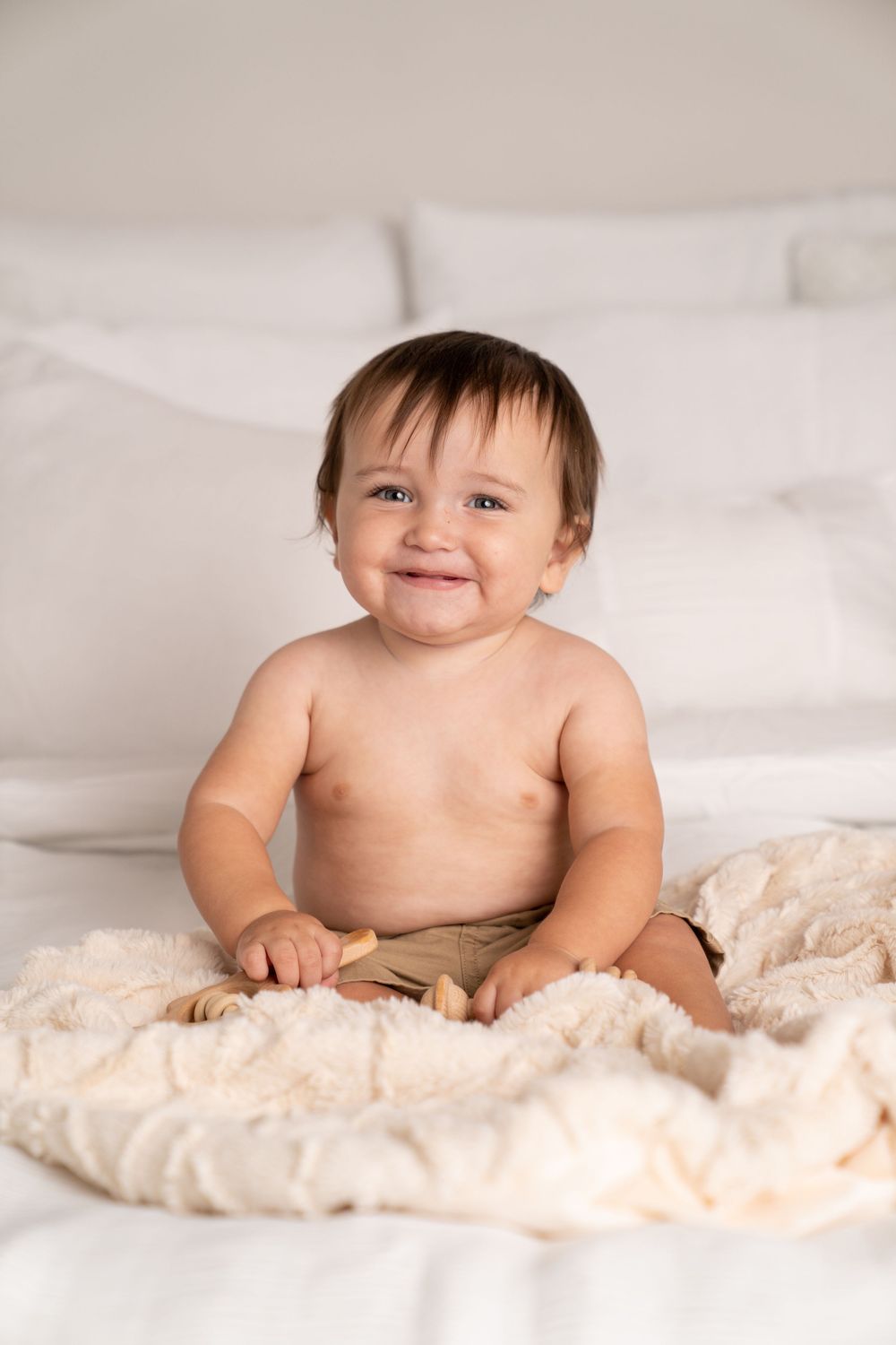 Smiling baby sitting on a white bed with a cream blanket, shirtless with dark hair and a joyful expression.