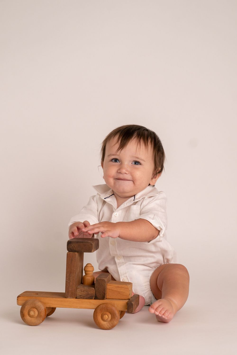Smiling baby in white outfit playing with a wooden toy truck on a light background.