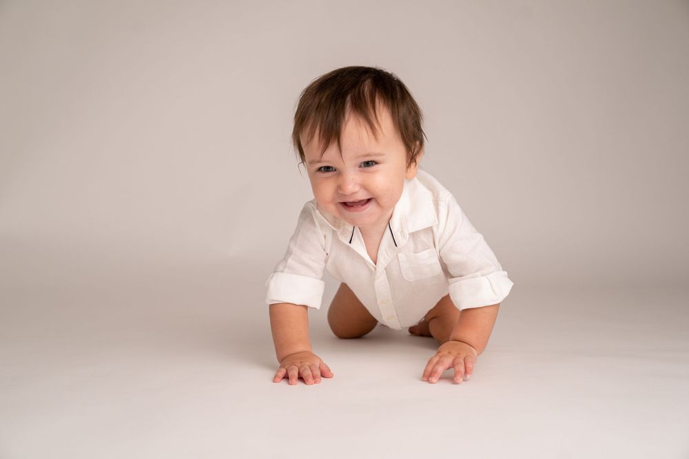 Smiling baby in white onesie crawling on white floor, cheerful infant portrait photography studio session.