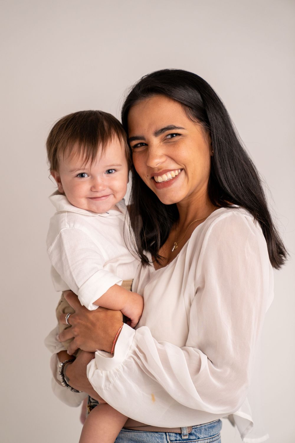 Smiling woman in white blouse holding a happy baby in white outfit against a light gray background.